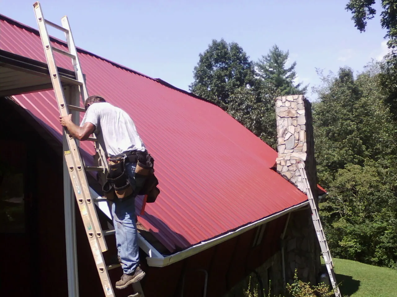 Roofer on a ladder beside a red metal roof and stone chimney — Soffit & Fascia Repair by Roofing Brothers in Hughson